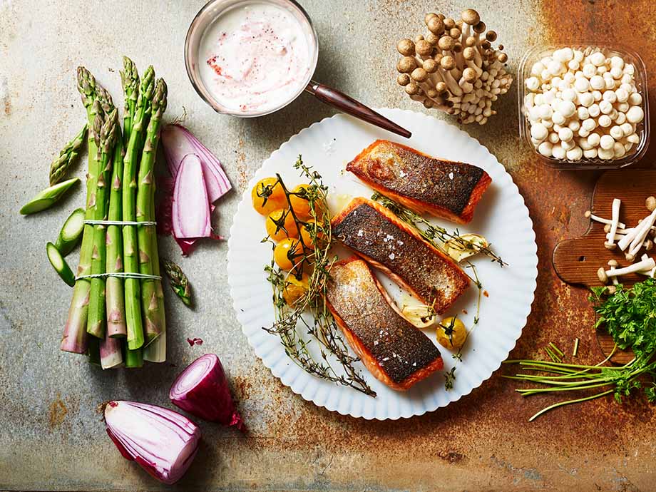 3 gebratene Lachsfilet Stücke auf weißem Teller. Als Dekoration liegen auf dem Teller noch orangene Cherrytomaten. Um den Teller herum liegen auf dem Tisch noch links grüner Spargel und Schalotte und rechts diverse Pilzsorten.